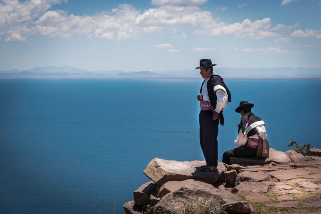 Two men in traditional attire stand on a cliff overlooking a serene ocean under a bright sky.